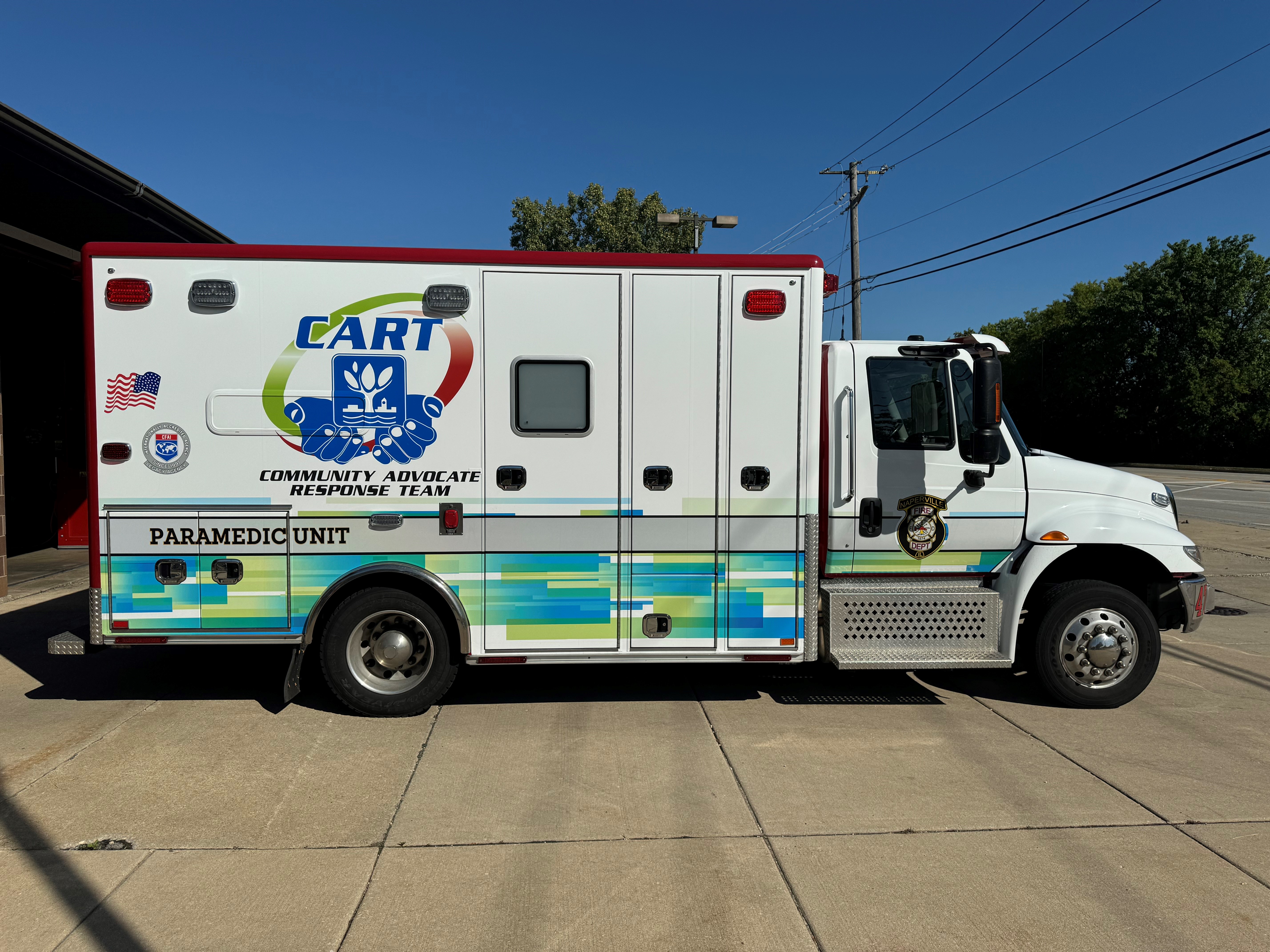 Community Advocate Response Team ambulance car. Car is white, with blue and green accents, and on its side is an image of the Naperville logo cradled by two hands. 