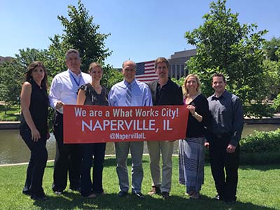 Naperville staff members hold a "What Works Cities" banner while standing on the riverwalk with the Municipal Center in the background