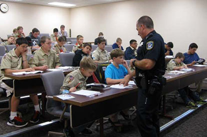 an officer teaches uniformed scouts, who are seated at long tables in a classroom setting