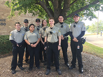 A group of uniformed police cadets stand under a tree in front of the Naperville Police Department