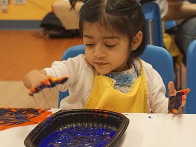 A child with brown hair and a yellow apron uses blue finger paint to make a hand print.
