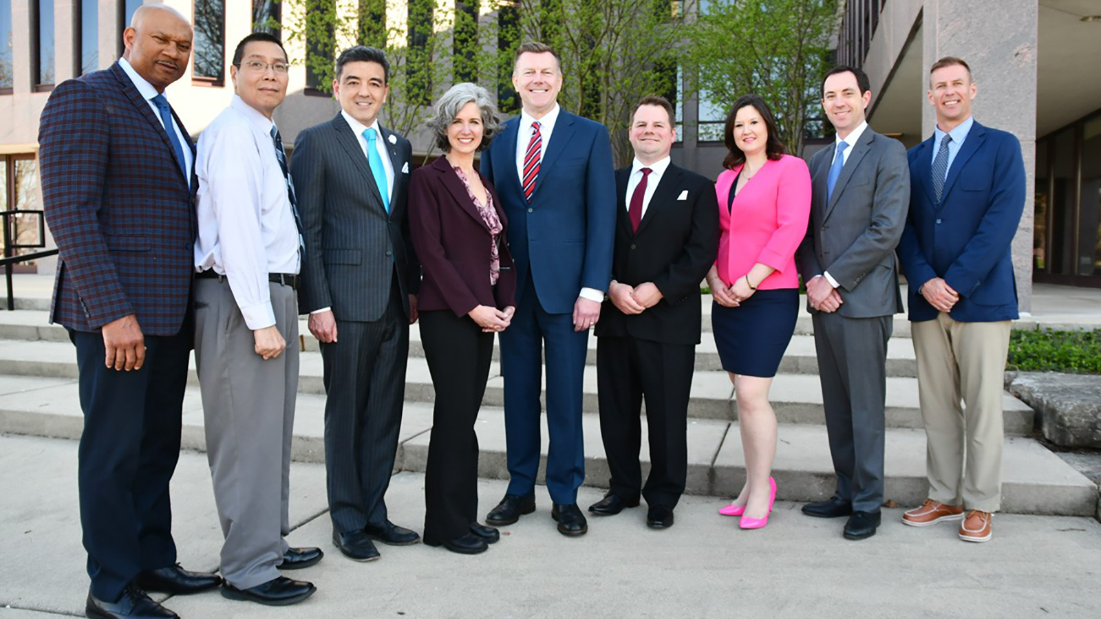 Mayor Scott Wehrli and eight Naperville City Council members stand together before the Municipal Center, along the riverwalk