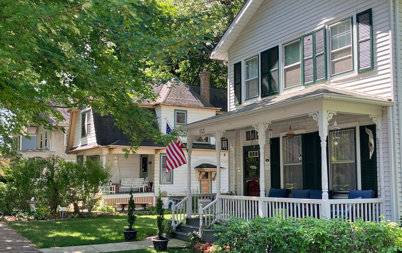 A white house with green shutters and a large front porch in Naperville's historic district displays an American flag.