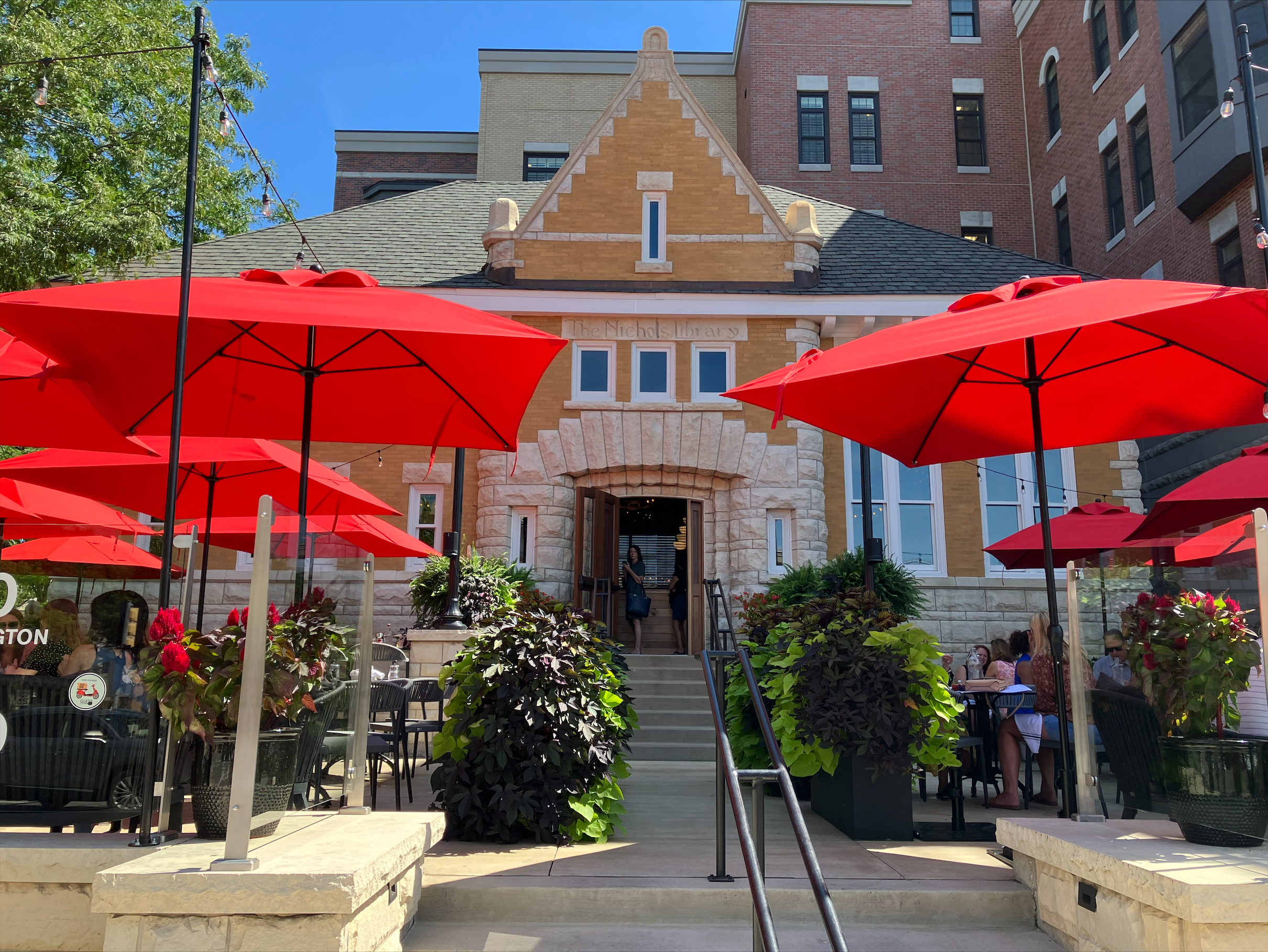 a limestone building, formerly a library, repurposed as a restaurant with tables and red umbrellas in front