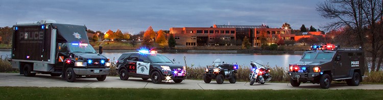 Police Department vehicles lined up with lights on in front of Lake Osborn and the Naperville Police Department in the background